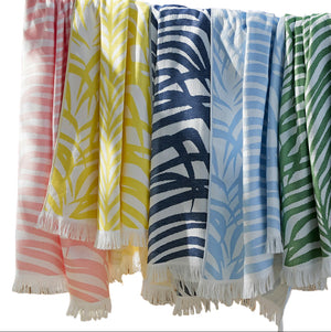 Five towels from the Matouk Zebra Beach Towel Collection in pastel pink, yellow, navy blue, light blue, and green with white tropical leaf patterns hang side by side on a line.