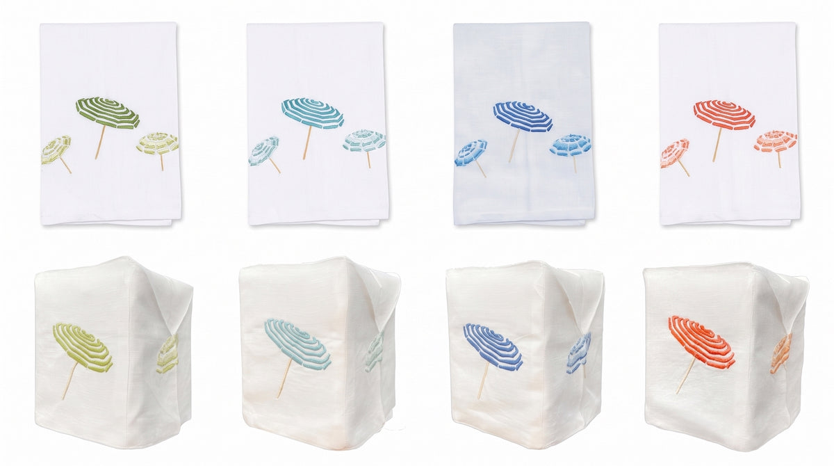 Eight white Italian linens from Haute Home’s Beach Umbrella Bath Collection: four flat linens with embroidered umbrellas in green, blue, or red; and four premium linen tissue box covers, each featuring matching umbrella embroidery.