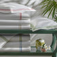 Neatly folded white sheets from the Matouk Amara Triple Pearl Bedding Collection, featuring a delicate triple pearl embroidered design along the edges, are stacked on a green shelf beside two pillows, a small vase, and a leafy plant in the background.