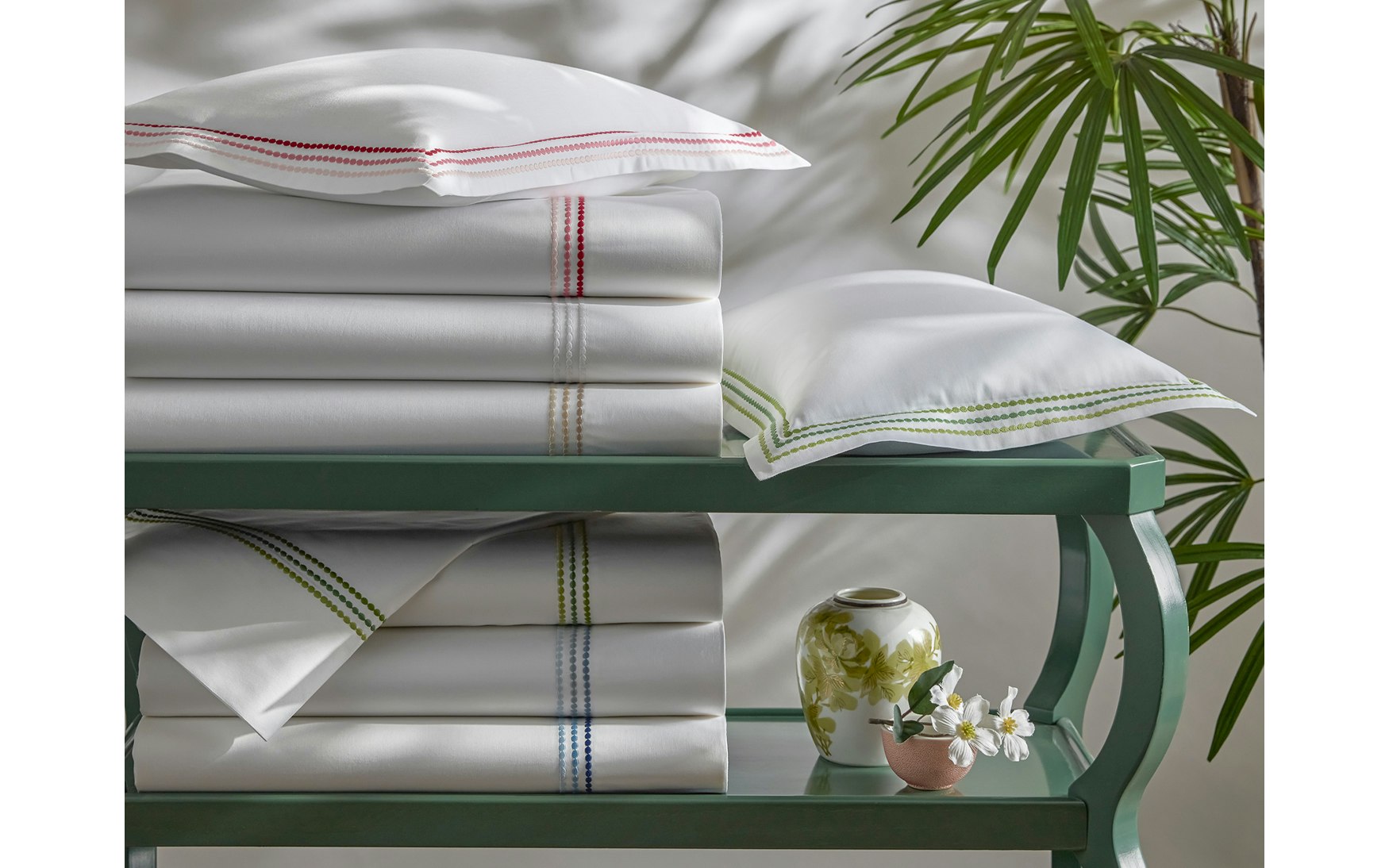 Neatly folded white sheets from the Matouk Amara Triple Pearl Bedding Collection, featuring a delicate triple pearl embroidered design along the edges, are stacked on a green shelf beside two pillows, a small vase, and a leafy plant in the background.