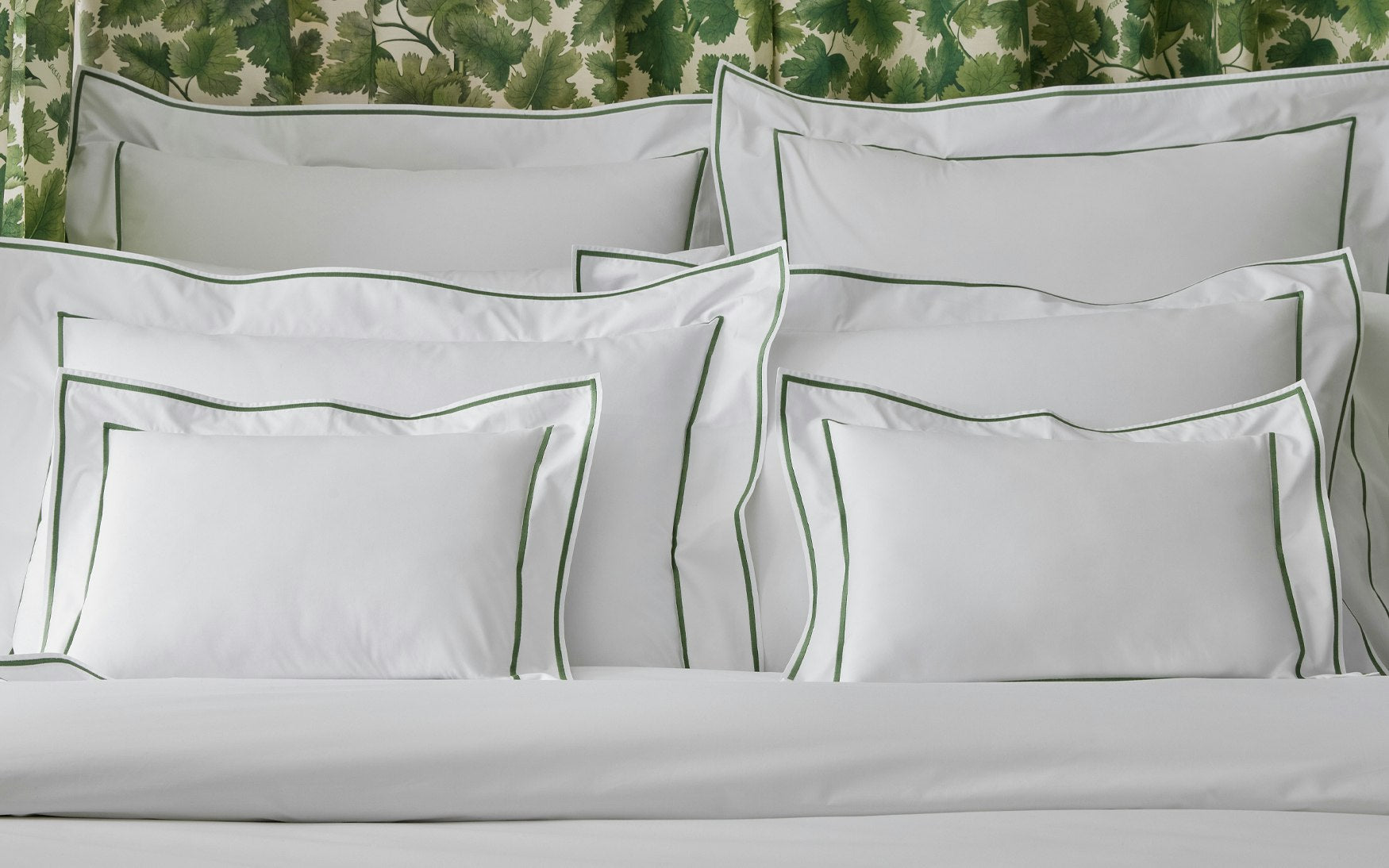 A neatly arranged bed with several white pillows featuring green trim from the Matouk Ansonia Bedding Collection, Palm, crafted in luxury Egyptian cotton percale, set against leafy green patterned wallpaper.