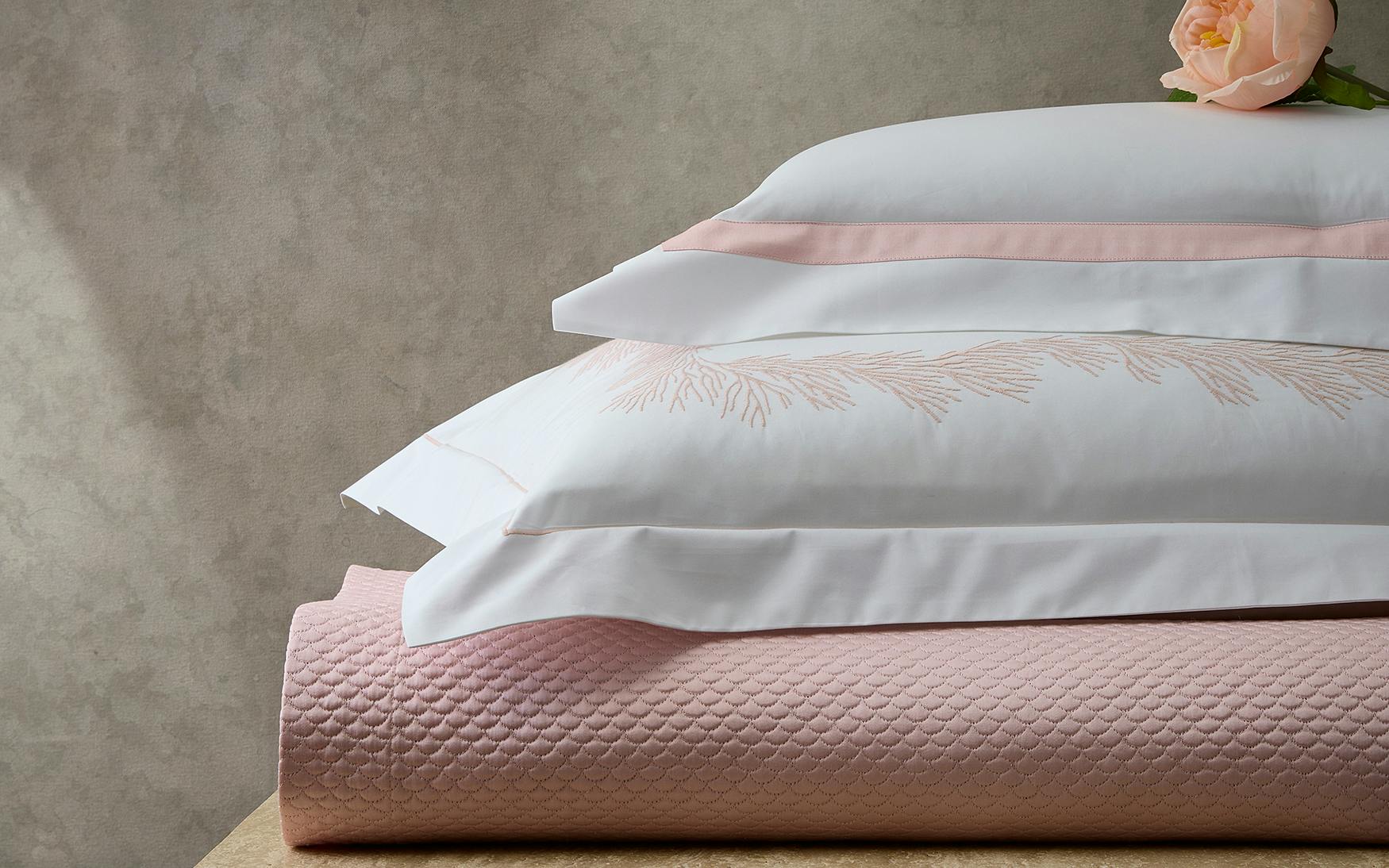 Two white pillows with embroidered patterns and pink trim from the Matouk Atoll Bedding Collection in Blush rest atop a folded, feathery coral-textured blanket, adorned with a peach-colored rose.