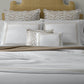 A neatly made bed with white and beige Astor Braid Matelassé Bedding Collection in Dune by Matouk, decorative pillows, a plant on the left nightstand, and three candles on the right nightstand.