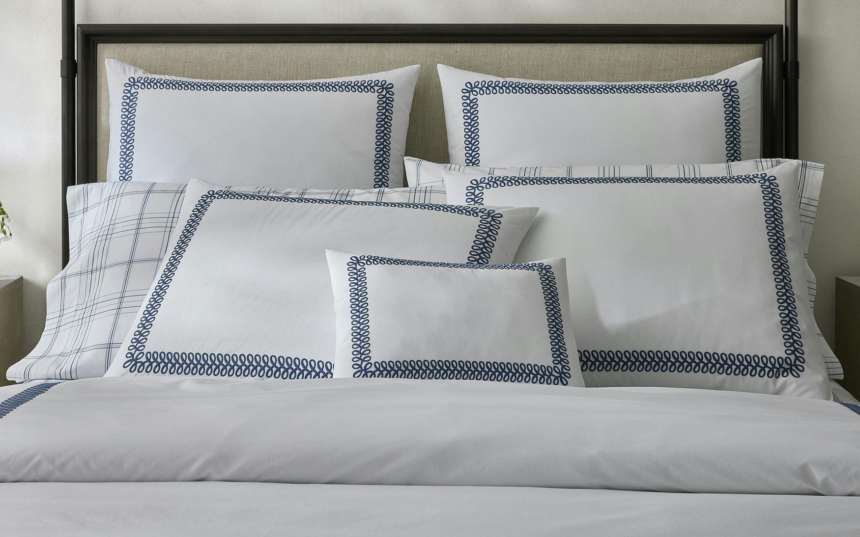 A neatly made bed showcases five white pillows featuring an indigo decorative border and a matching duvet from the Matouk Astor Braid Bedding Collection, exuding elegance.