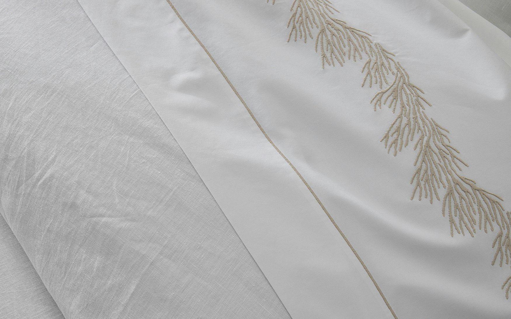 Close-up of white Matouk Atoll Bedding Collection cotton percale bed linens, one adorned with a beige, embroidered branch motif and the other displaying a plain, textured surface. The luxurious 520 thread count guarantees exceptional softness and durability.