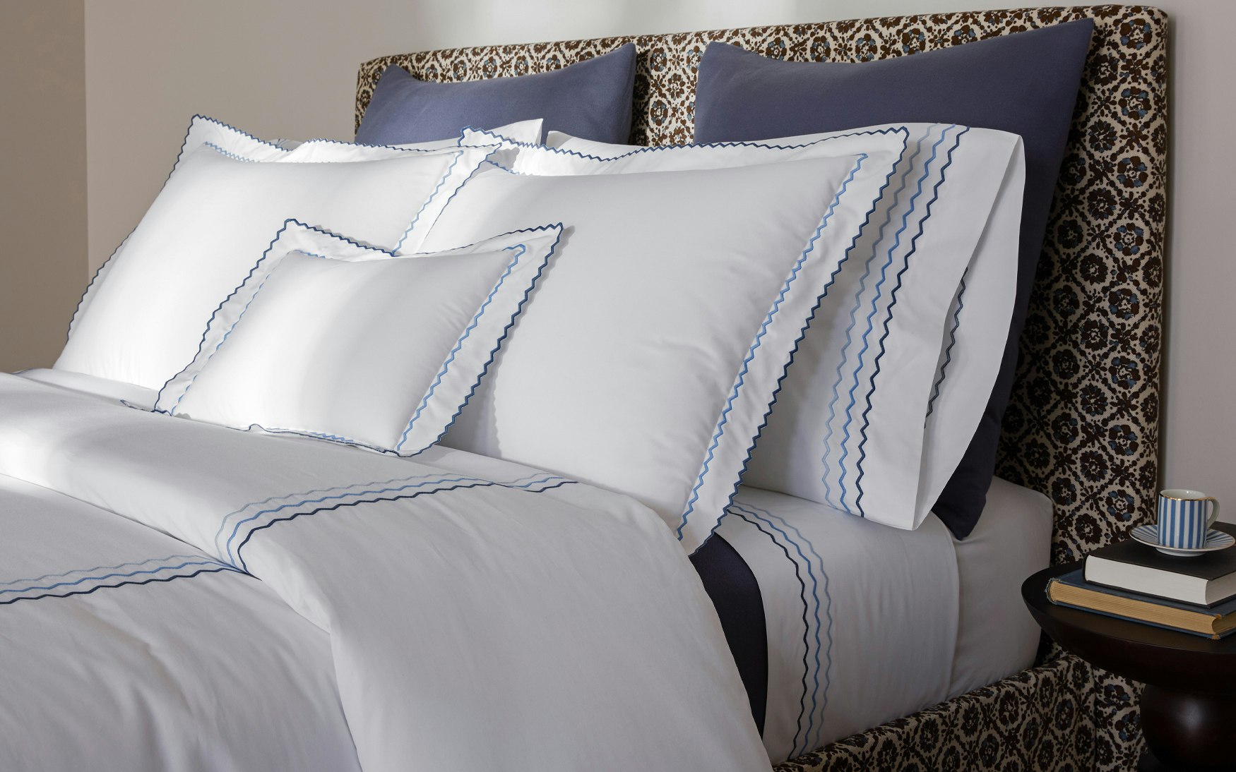 The Axel Bedding Collection in Azure by Matouk features white long-staple cotton linens with blue scalloped edges, dark blue accent pillows, and a patterned headboard. A small round table with books and a striped cup sits beside the bed.