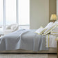 A modern bedroom features a neatly made bed with Matouk's Alba Bedding Collection in Pool, decorative pillows, bedside tables with lamps, a round chair, large windows, and a city view.