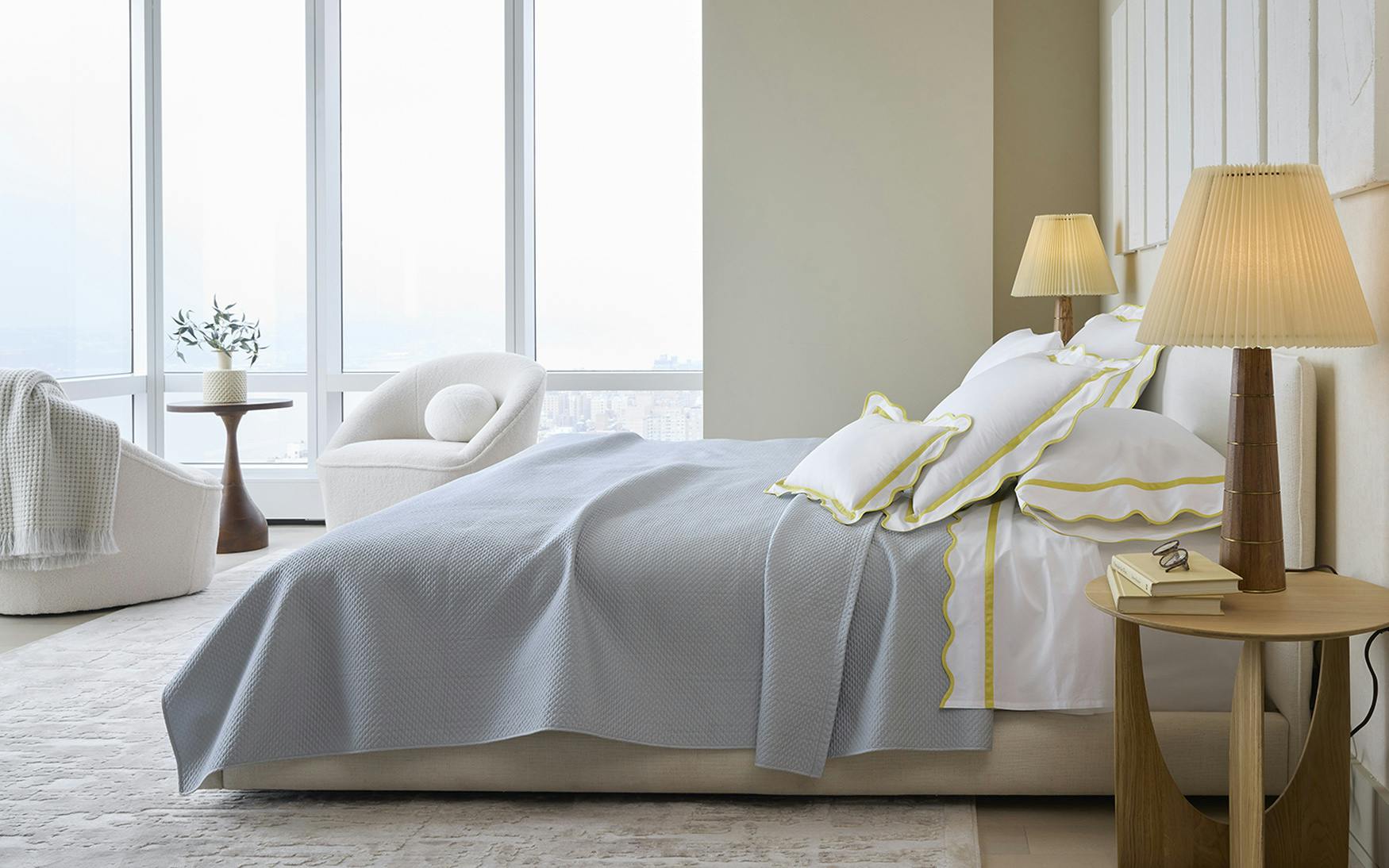 A modern bedroom features a neatly made bed with Matouk's Alba Bedding Collection in Pool, decorative pillows, bedside tables with lamps, a round chair, large windows, and a city view.