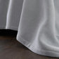 The Costa Mesa Blanket Collection by Matouk features a light gray, heavyweight, soft-knit blanket that drapes over the bed’s edge, reaching down to a dark wooden floor.