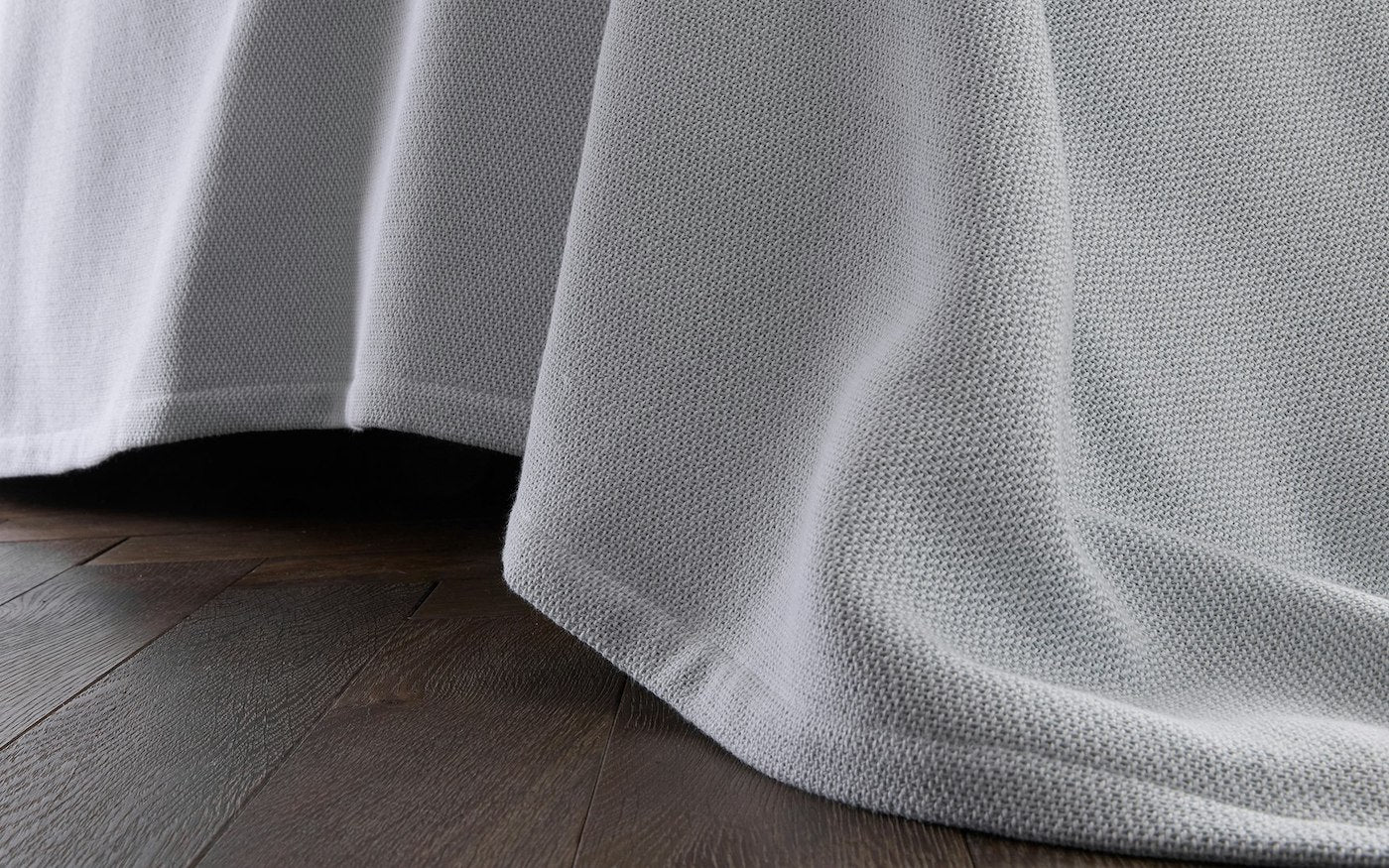 The Costa Mesa Blanket Collection by Matouk features a light gray, heavyweight, soft-knit blanket that drapes over the bed’s edge, reaching down to a dark wooden floor.