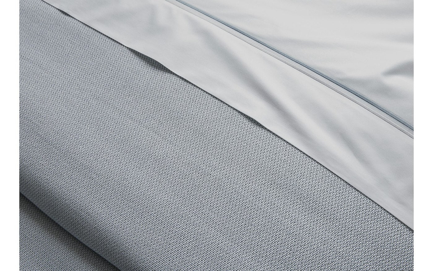 Close-up of the Matouk Costa Mesa Blanket Collection in light gray, highlighting its soft knitted texture and a smooth contrasting sheet, with visible seams and folds for added detail.