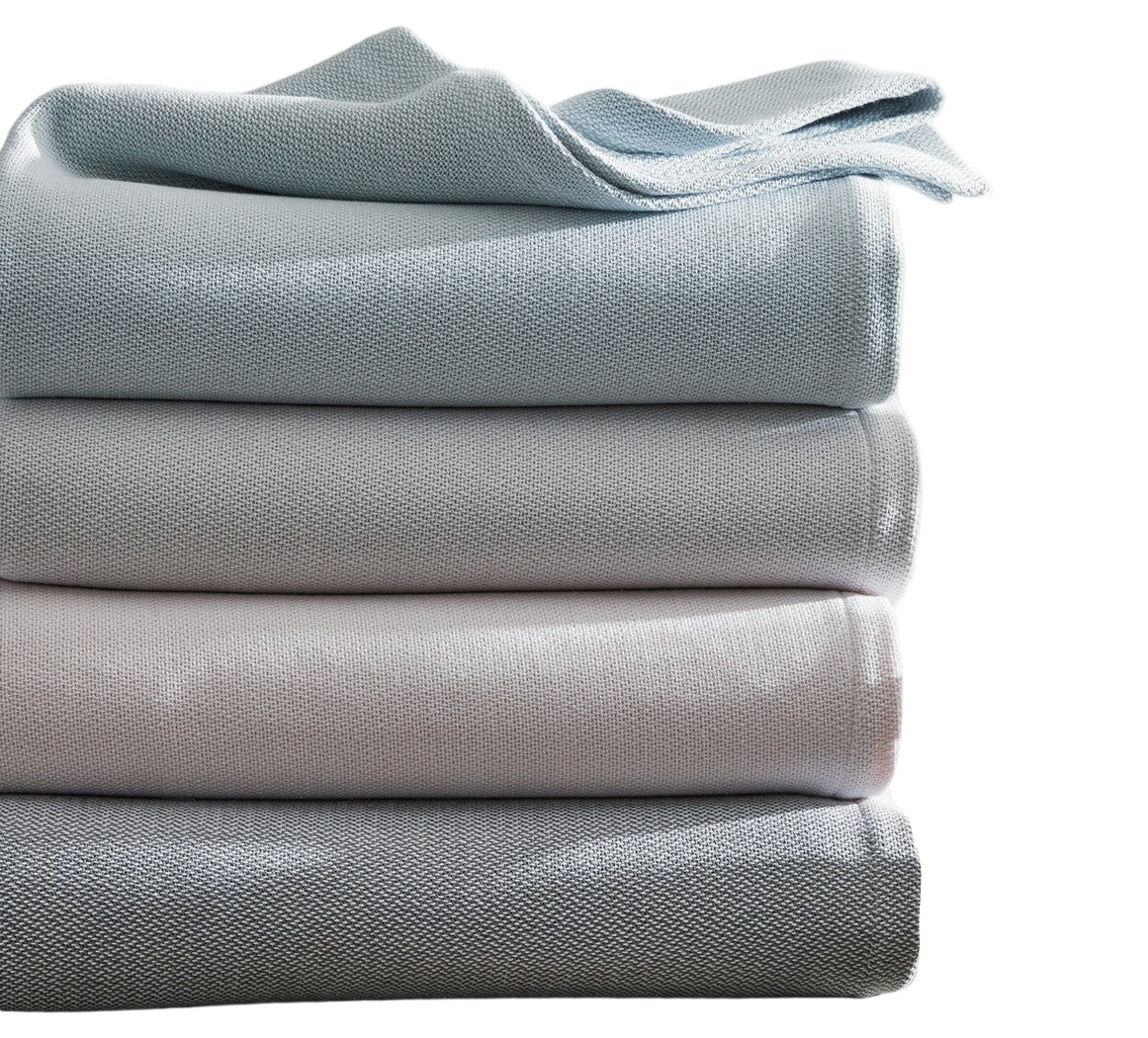 The Costa Mesa Blanket Collection by Matouk features four heavyweight, textured knit blankets in grey and light blue, neatly folded and displayed against a white background.