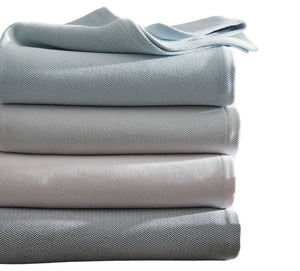 The Costa Mesa Blanket Collection by Matouk features four heavyweight, textured knit blankets in grey and light blue, neatly folded and displayed against a white background.