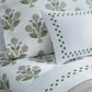 Neatly arranged on the bed are white bedsheets and pillowcases from the Dalila Bedding Collection by Matouk, featuring green and gray floral patterns reminiscent of a vibrant Schumacher block print. One pillow is styled with a Luca percale cover by Matouk, accented with a green geometric border design.
