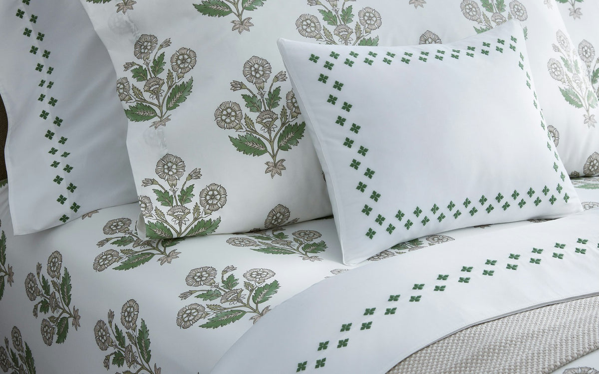 Neatly arranged on the bed are white bedsheets and pillowcases from the Dalila Bedding Collection by Matouk, featuring green and gray floral patterns reminiscent of a vibrant Schumacher block print. One pillow is styled with a Luca percale cover by Matouk, accented with a green geometric border design.