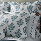 A neatly made bed with white pillows and bedding from the Dalila Bedding Collection by Matouk, featuring a vibrant floral blue and green pattern reminiscent of a block print, next to a vase with red branches.