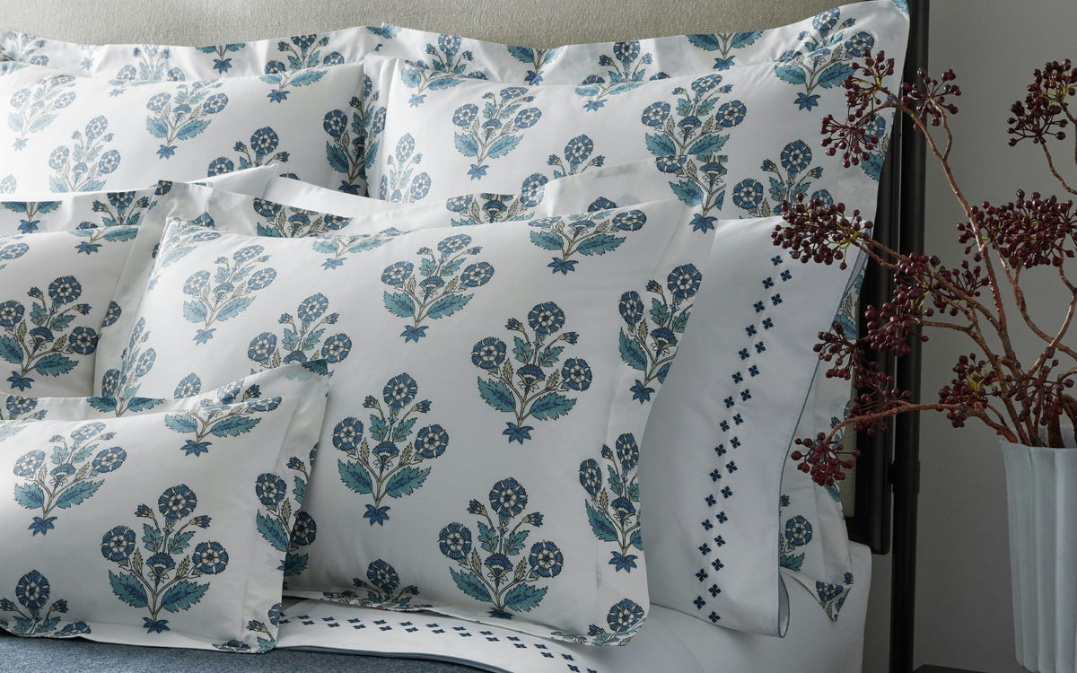 A neatly made bed with white pillows and bedding from the Dalila Bedding Collection by Matouk, featuring a vibrant floral blue and green pattern reminiscent of a block print, next to a vase with red branches.
