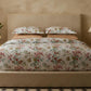 A bed dressed in Matouk’s Del Tesoro Bedding Collection, Coral, conjures an oceanic fantasy and is flanked by wooden nightstands with a potted plant, lamp, and glass bottles in a neutral-toned room.