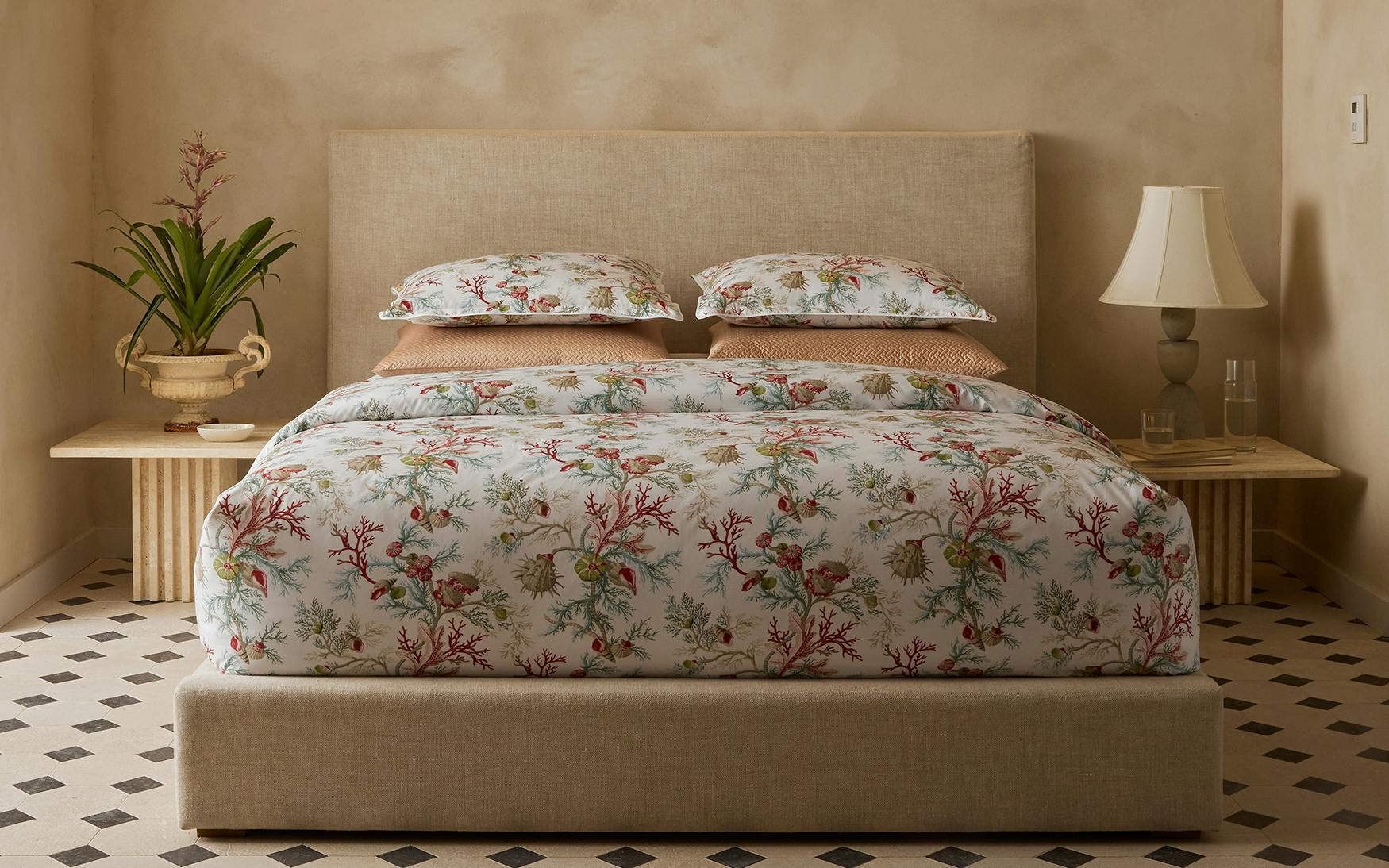 A bed dressed in Matouk’s Del Tesoro Bedding Collection, Coral, conjures an oceanic fantasy and is flanked by wooden nightstands with a potted plant, lamp, and glass bottles in a neutral-toned room.