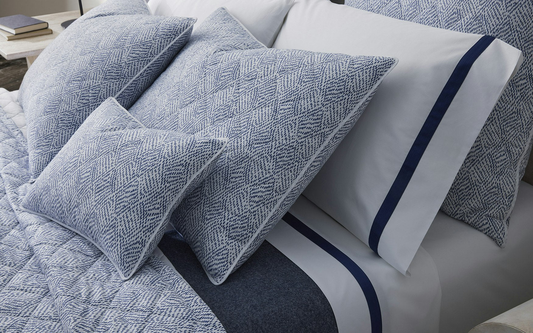 A neatly made bed featuring Matouk's Lowell Bedding Collection in Navy, with blue and white patterned pillows, a comforter, and crisp Egyptian cotton percale sheets accented by a navy sateen tape border.