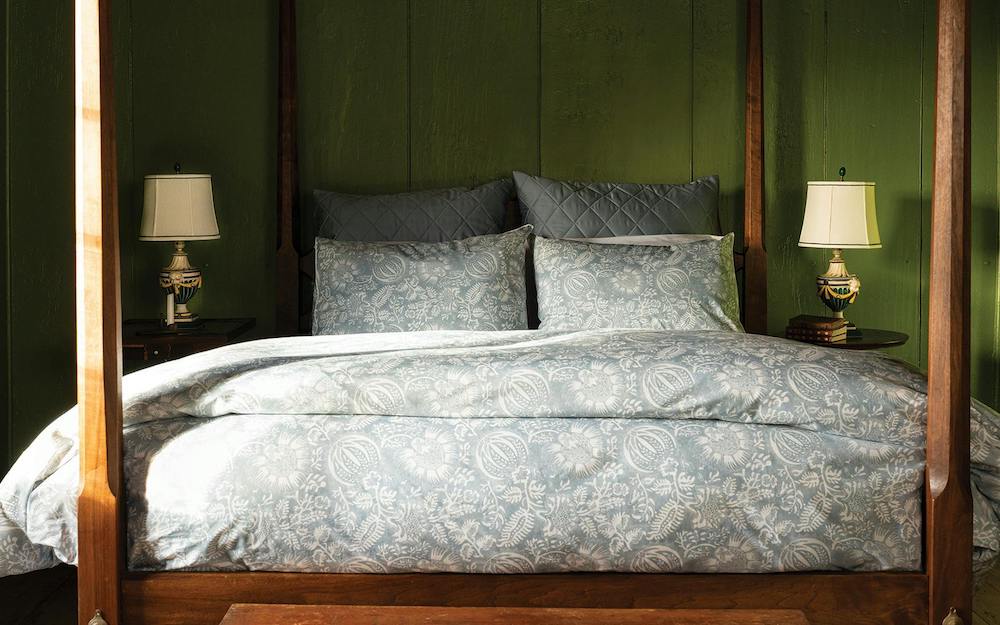 A wooden four-poster bed with Matouk's Granada Bedding Collection in Hazy Blue is set against a green wall, flanked by two small tables with matching lamps.