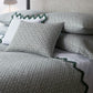 A neatly made bed features the Laksha Bedding Collection by Matouk—patterned white and green bedding, including pillows and shams crafted from luxurious Luca percale—set against a light upholstered headboard.