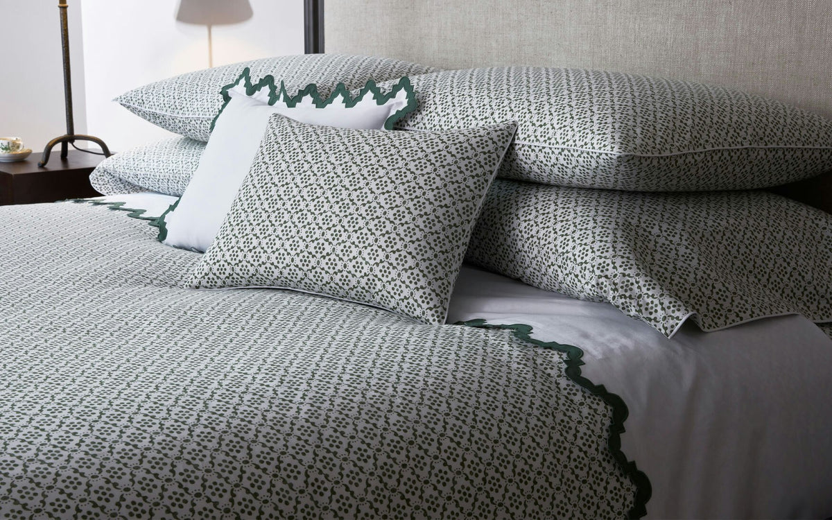 A neatly made bed features the Laksha Bedding Collection by Matouk—patterned white and green bedding, including pillows and shams crafted from luxurious Luca percale—set against a light upholstered headboard.
