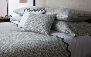 A neatly made bed features the Laksha Bedding Collection by Matouk—patterned white and green bedding, including pillows and shams crafted from luxurious Luca percale—set against a light upholstered headboard.