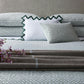 A neatly made bed featuring the Matouk Laksha Bedding Collection, accented with a brown plaid blanket, three pillows, and a sprig of purple flowers placed on top.