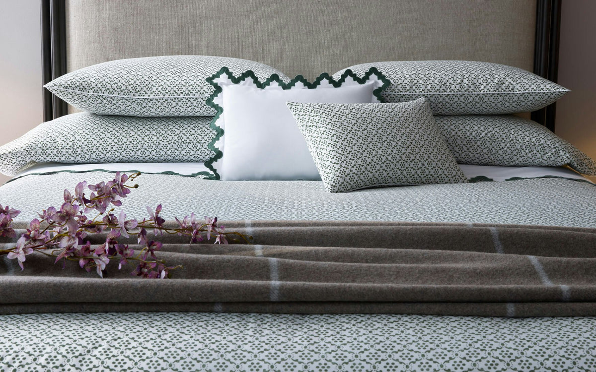 A neatly made bed featuring the Matouk Laksha Bedding Collection, accented with a brown plaid blanket, three pillows, and a sprig of purple flowers placed on top.