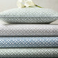 A stack of three folded bed sheets from the Matouk Laksha Bedding Collection—in brown, green, and blue, featuring traditional Indian block print-inspired patterns on soft percale—rests together with a coordinating pillow and two white calla lilies on top, set against a light-colored wall.