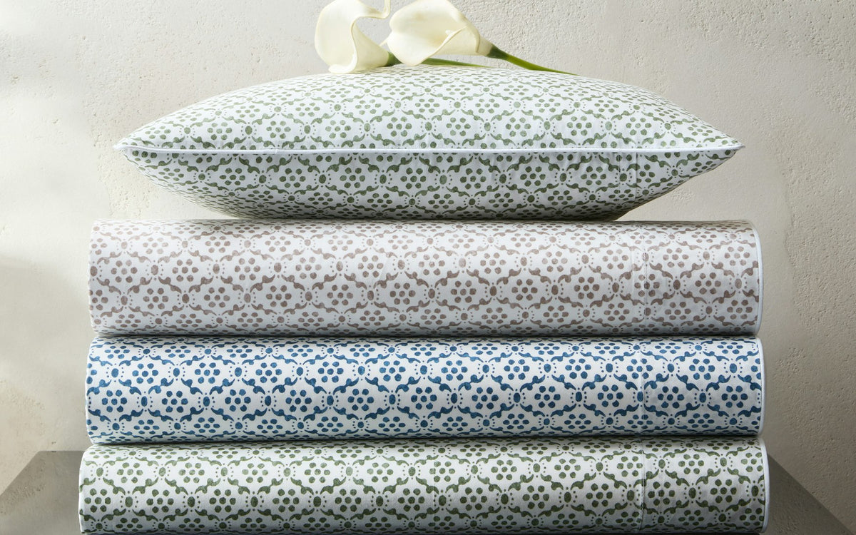 A stack of three folded bed sheets from the Matouk Laksha Bedding Collection—in brown, green, and blue, featuring traditional Indian block print-inspired patterns on soft percale—rests together with a coordinating pillow and two white calla lilies on top, set against a light-colored wall.