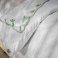 Close-up of Matouk’s Marcella Bedding Collection in Clover, featuring green scalloped floral embroidery, draped elegantly over the side of a bed on a patterned floor.