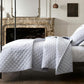 A neatly made bed with Matouk’s Bergamo Hemstitch Bedding Collection in white stands before a marble fireplace in a modern, minimal bedroom.
