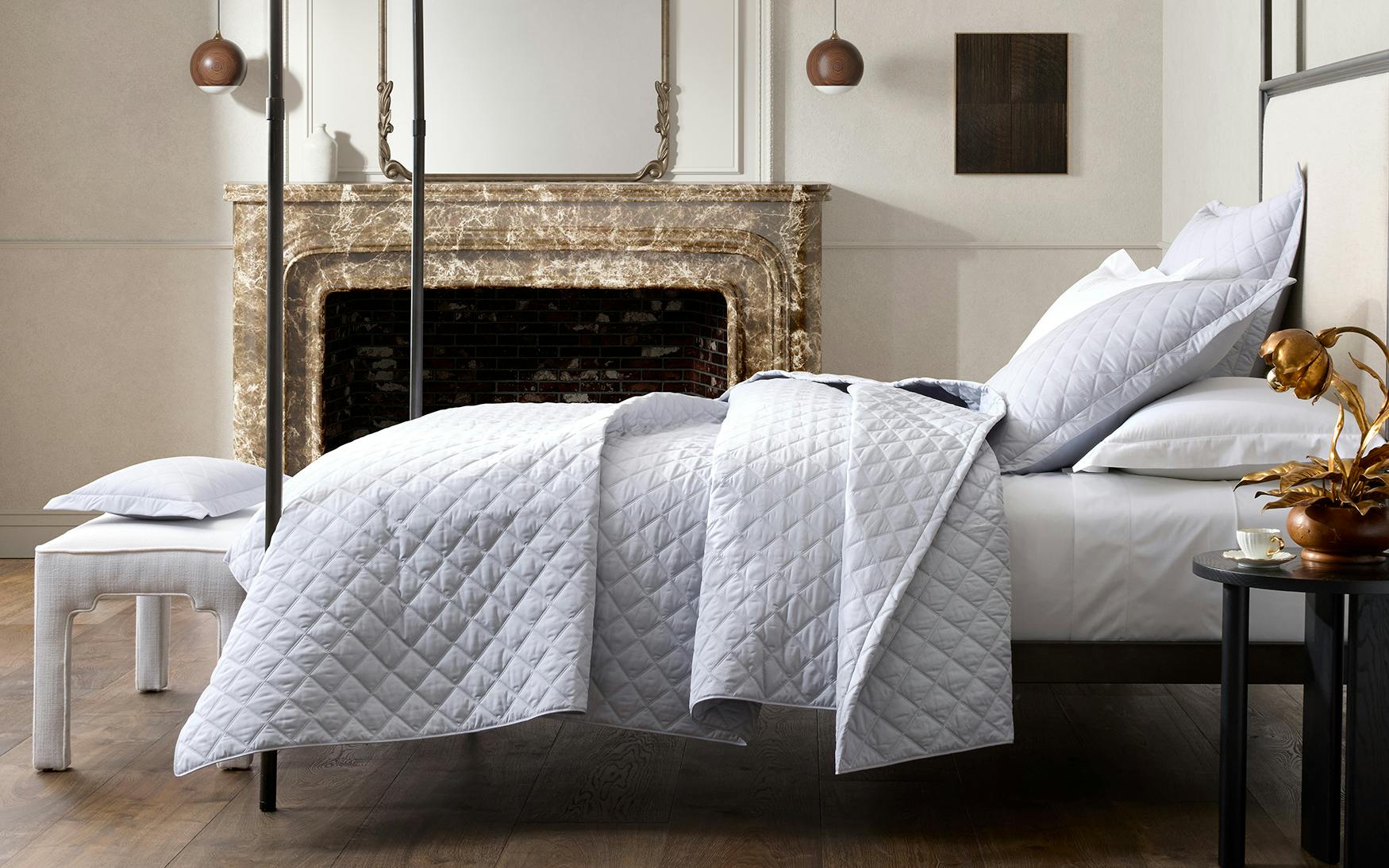 A neatly made bed with Matouk’s Bergamo Hemstitch Bedding Collection in white stands before a marble fireplace in a modern, minimal bedroom.