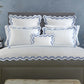 A neatly made bed features the Matouk Mirasol Bedding Collection in Azure, flaunting scalloped-edged white and blue luxury bedding made from 600 thread count Egyptian cotton percale, flanked by a nightstand holding a vase of flowers and books, set against a light blue wall.