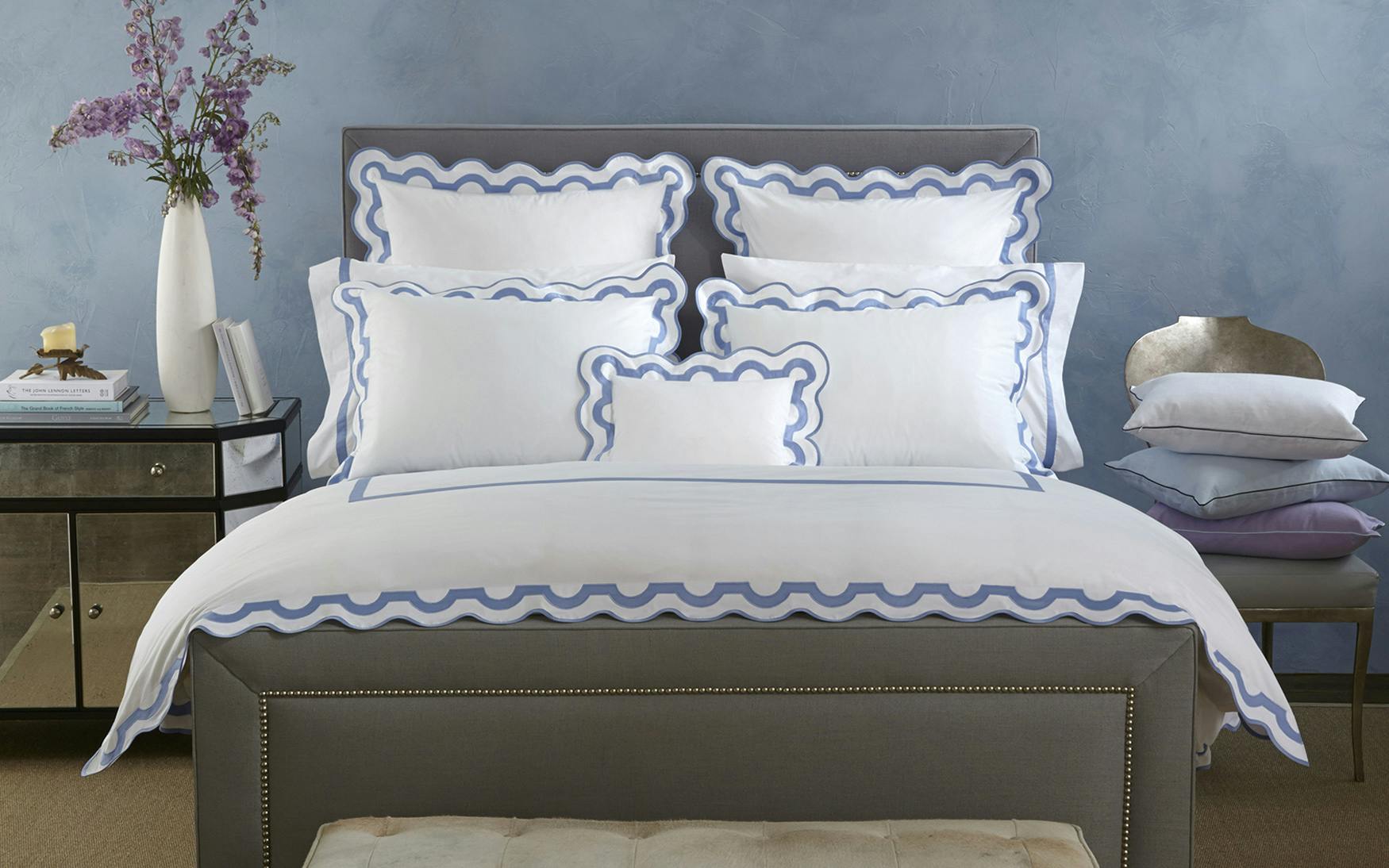 A neatly made bed features the Matouk Mirasol Bedding Collection in Azure, flaunting scalloped-edged white and blue luxury bedding made from 600 thread count Egyptian cotton percale, flanked by a nightstand holding a vase of flowers and books, set against a light blue wall.