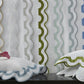 Vibrant white pillows with ruffled blue and green edges from the Matouk Mirasol Bedding Collection sit in front of sheer curtains, each edged with large, wavy stripes in pink, tan, green, blue, and gray. Crafted from luxurious 600 thread-count Egyptian cotton percale, this ensemble adds both comfort and lively color to any room.