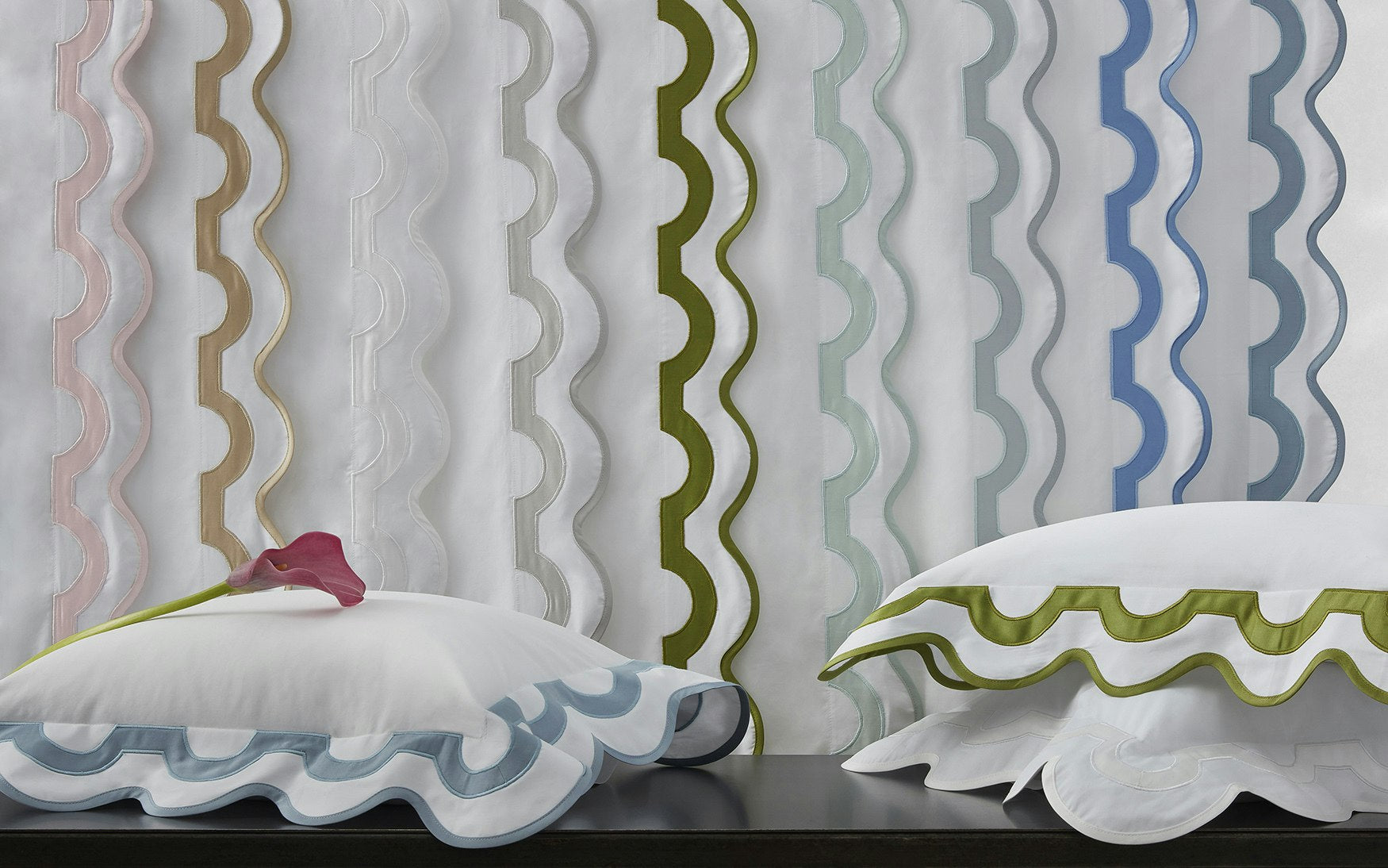 Vibrant white pillows with ruffled blue and green edges from the Matouk Mirasol Bedding Collection sit in front of sheer curtains, each edged with large, wavy stripes in pink, tan, green, blue, and gray. Crafted from luxurious 600 thread-count Egyptian cotton percale, this ensemble adds both comfort and lively color to any room.