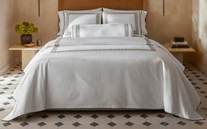 A minimalist room features a neatly made bed with white linens, gray Greek key borders, and the Matouk Ophelia Mocha Bolster w/Insert, set atop hexagonal tile flooring beside a small table holding a vase and candle.