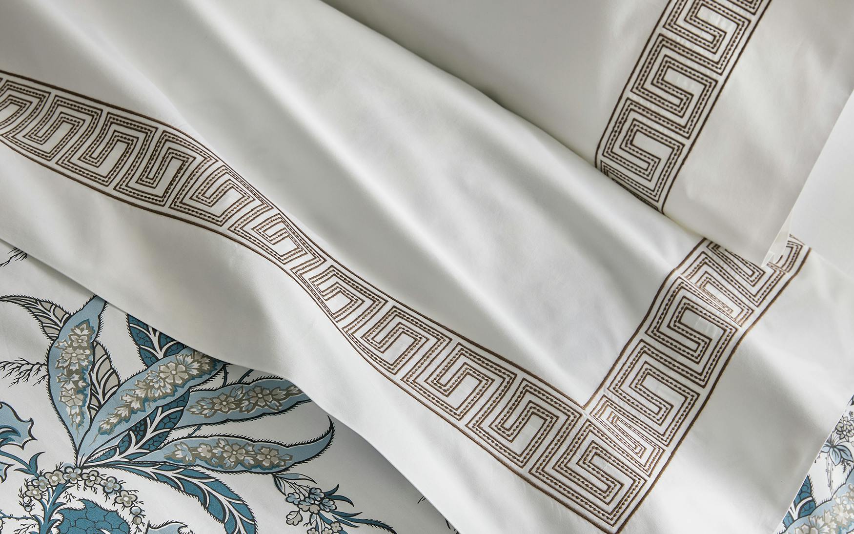 Matouk’s Ophelia Bedding Collection, Mocha, features white sheets and pillowcases with a brown Greek key embroidery layered over blue and white florals. Crafted with care in 520 thread count, this set adds elegance to any bedroom.