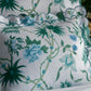 A decorative pillow with a scalloped edge and green-blue floral pattern rests on Matouk's Palmyra Bedding Collection in Emerald, with a stack of books and a potted plant in the background.