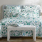 A neatly made bed features the Matouk Palmyra Bedding Collection in Emerald, accented by white and green palm fronds. Two lamps rest on side tables, with a bench positioned at the foot of the bed.