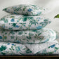 Four items from the Matouk Palmyra Bedding Collection in Emerald, featuring a green and blue tropical design, are stacked on a table beside a glass vase with green leaves and a small white cup.
