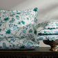 Four decorative pillows from Matouk's Palmyra Bedding Collection in Emerald, featuring blue and green botanical patterns with scalloped edges, are displayed on dark wood; three are stacked in a glass bowl.