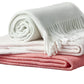 Three folded blankets from the Matouk Pezzo Throw Collection are stacked: a white fringed throw on top, light pink in the middle, and coral pink at the bottom—all made from soft, OEKO-TEX certified materials.