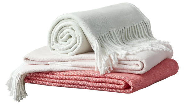 A cozy throw stack features the Matouk Pezzo Throw Collection: a white fringed blanket on top, a light pink brushed cotton one in the middle, and a coral gem at the bottom, each crafted by Matouk and OEKO-TEX Standard 100 certified for ultimate comfort.