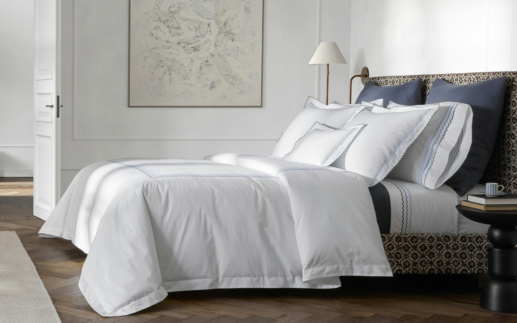 A modern bed dressed in Matouk's Axel Bedding Collection, Azure, with white and blue zigzag embroidery, navy accent pillows, a patterned headboard, and a bedside table with a cup in soft lighting.