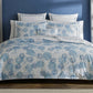 A bed dressed in Matouk’s Tillett x Matouk Chrysanthemum Bedding Collection in blue, flanked by two lamps on wooden nightstands, set against a blue wall with light gray curtains.
