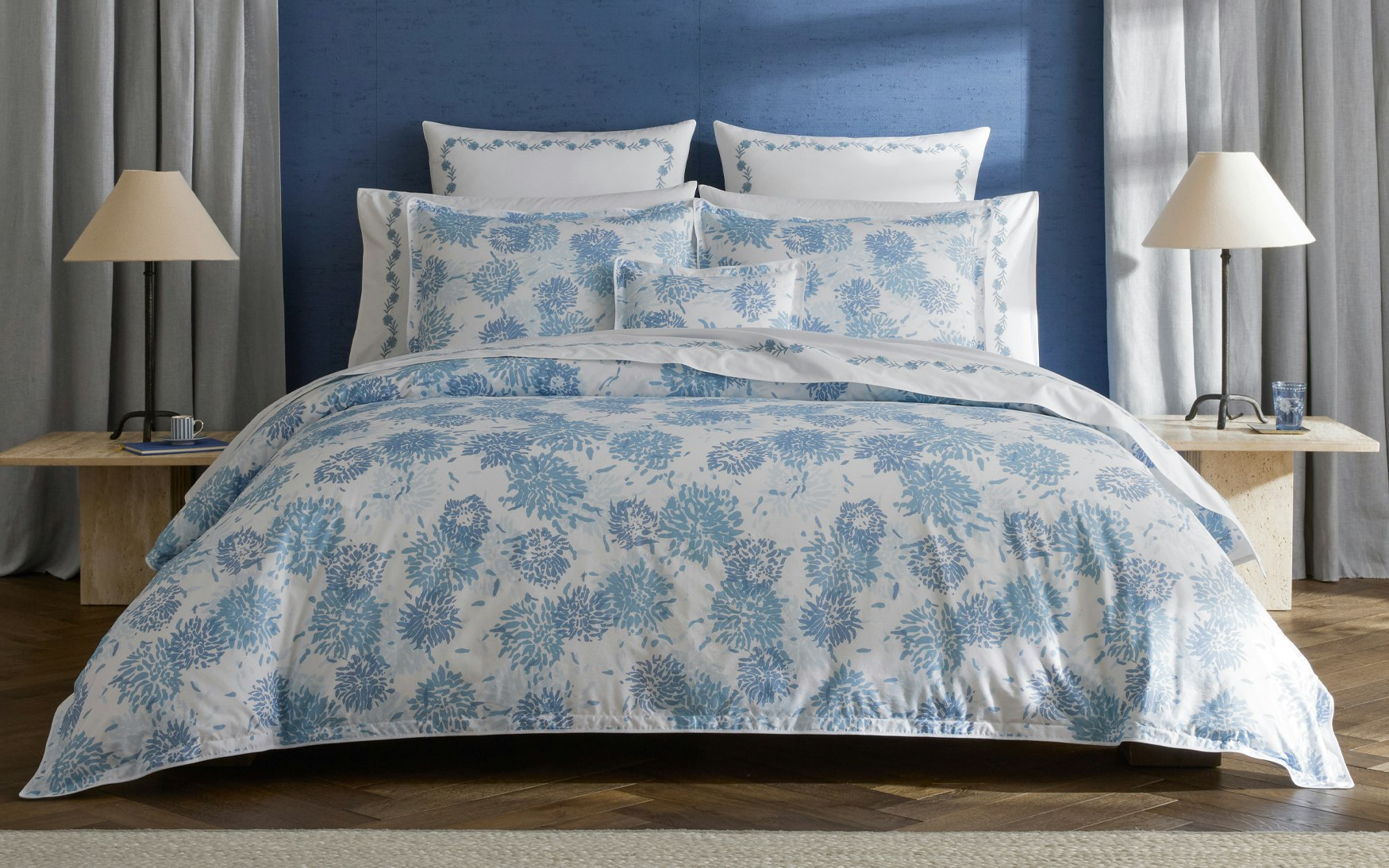 A bed dressed in Matouk’s Tillett x Matouk Chrysanthemum Bedding Collection in blue, flanked by two lamps on wooden nightstands, set against a blue wall with light gray curtains.